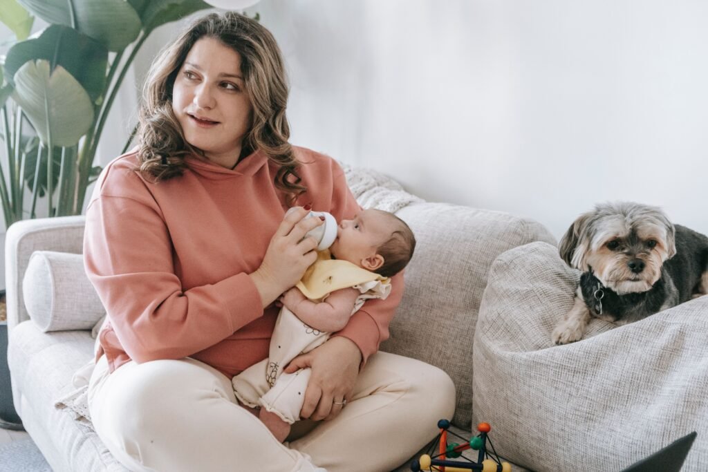 Caring mother looking away while feeding baby with bottle on soft sofa near hairy dog in light room at home
